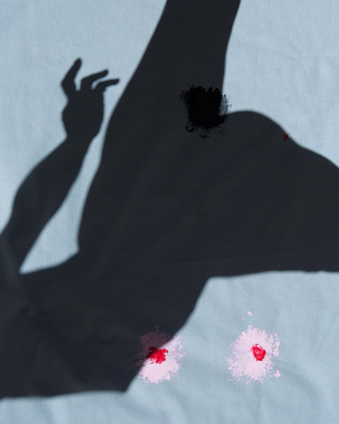 Abstract shadow of an arm and hand casting on off-white fabric, complemented by scattered red and pink paint splatters.