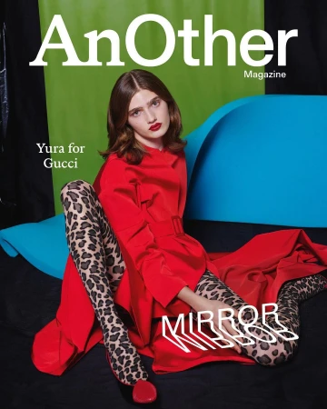 A female model wearing a vibrant red coat and leopard print tights seated on a vibrant blue and green studio set.
