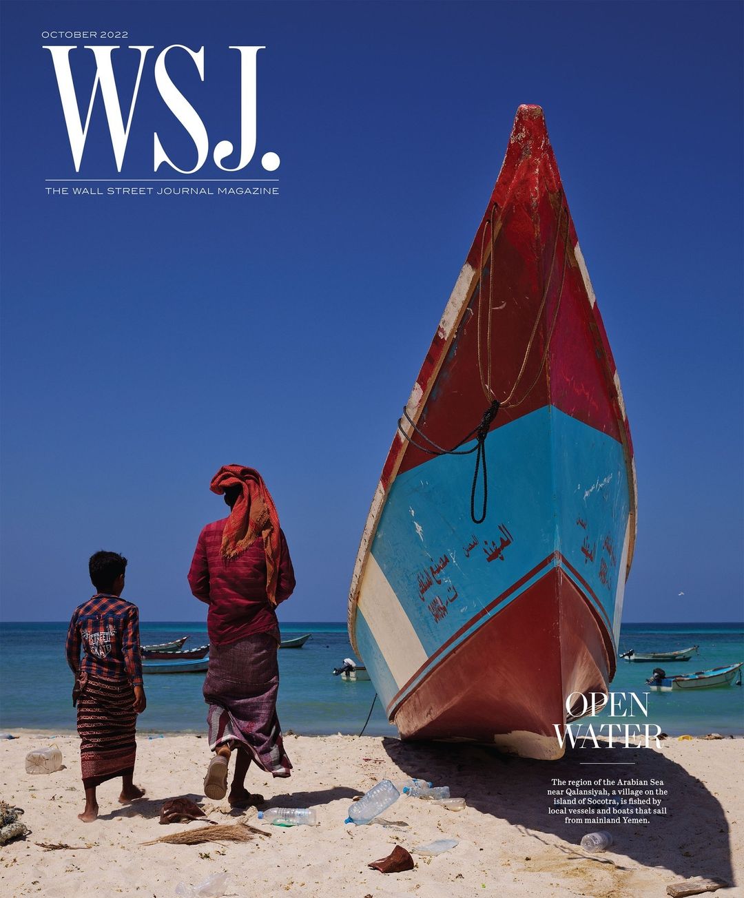 Magazine cover with a vibrant fishing boat on a sandy shore near two figures in traditional dress against a bright blue sky.