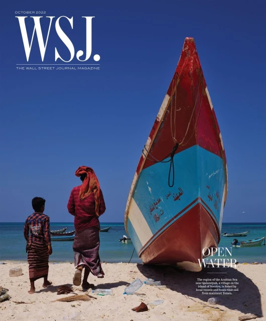 Magazine cover with a vibrant fishing boat on a sandy shore near two figures in traditional dress against a bright blue sky.