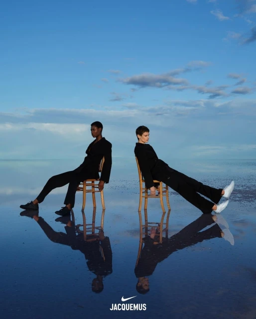 Two fashion models sit on chairs in a mirrored landscape reflecting the sky. The models are wearing black suits with athletic shoes.