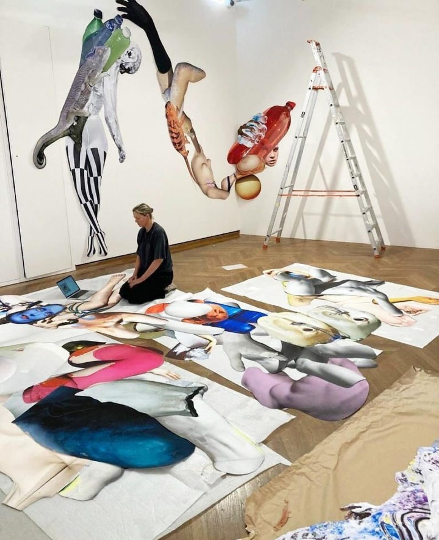 Woman kneeling among large-scale mixed media artworks, including paper cutouts, illustrations, and collage in a bright, white art studio.