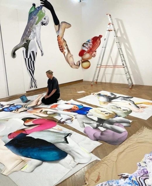 Woman kneeling among large-scale mixed media artworks, including paper cutouts, illustrations, and collage in a bright, white art studio.