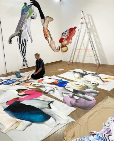 Woman kneeling among large-scale mixed media artworks, including paper cutouts, illustrations, and collage in a bright, white art studio.