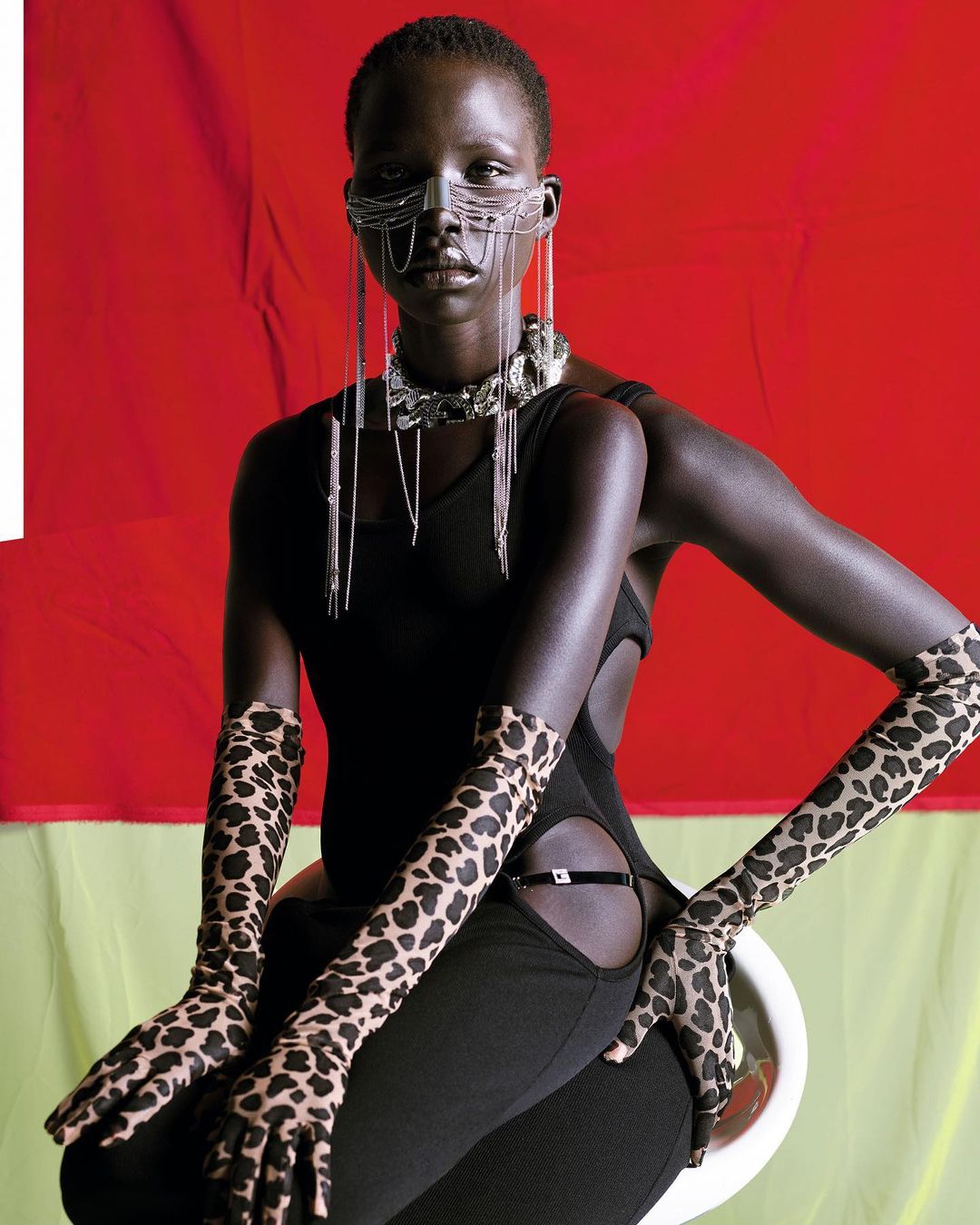 Woman with dark skin poses wearing black dress and animal print gloves with silver mask and necklace, set against red and yellow backdrop.