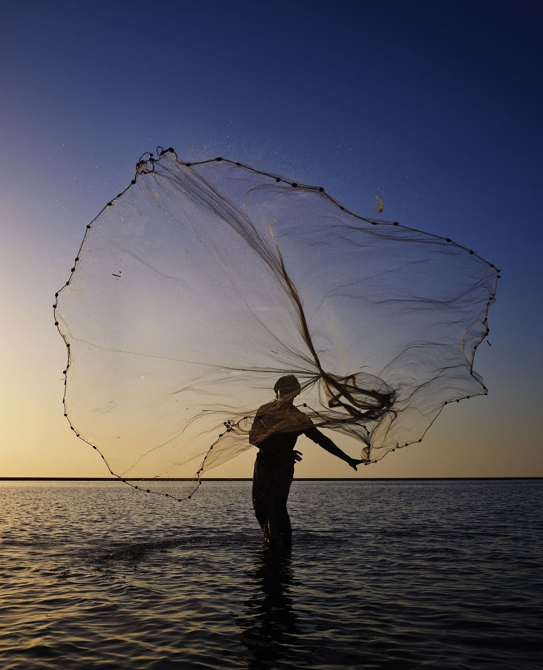 Fisherman casts a net into the sea at dusk, creating a circular pattern in the air against a gradient sky, shallow depth of field.
