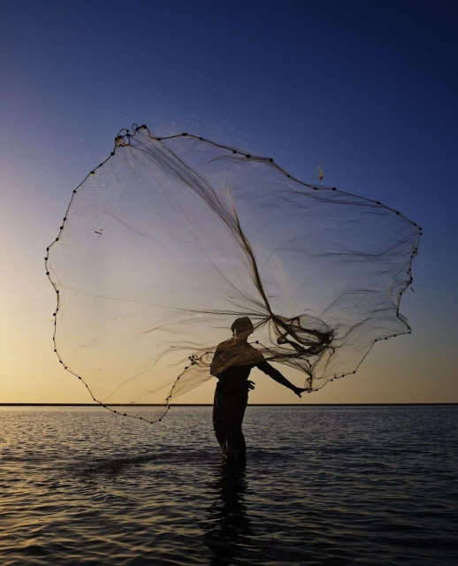 Fisherman casts a net into the sea at dusk, creating a circular pattern in the air against a gradient sky, shallow depth of field.