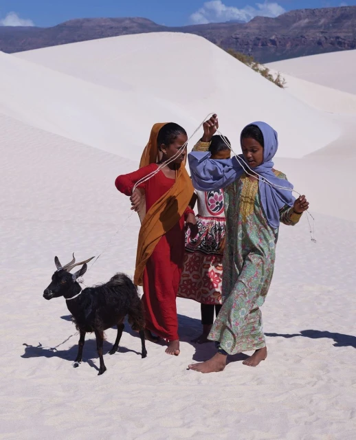 Three young girls in traditional clothing play with a black goat in a white sand dune landscape under a clear blue sky.