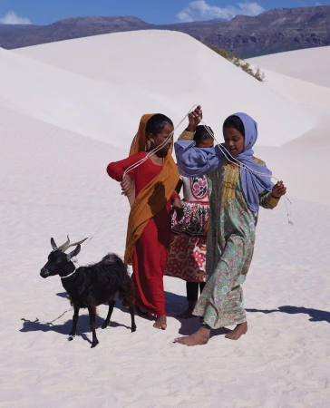 Three young girls in traditional clothing play with a black goat in a white sand dune landscape under a clear blue sky.