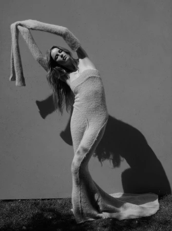 Model in a long textured knit dress with matching arm warmers arching back against a plain wall photographed by Viviane Sassen.
