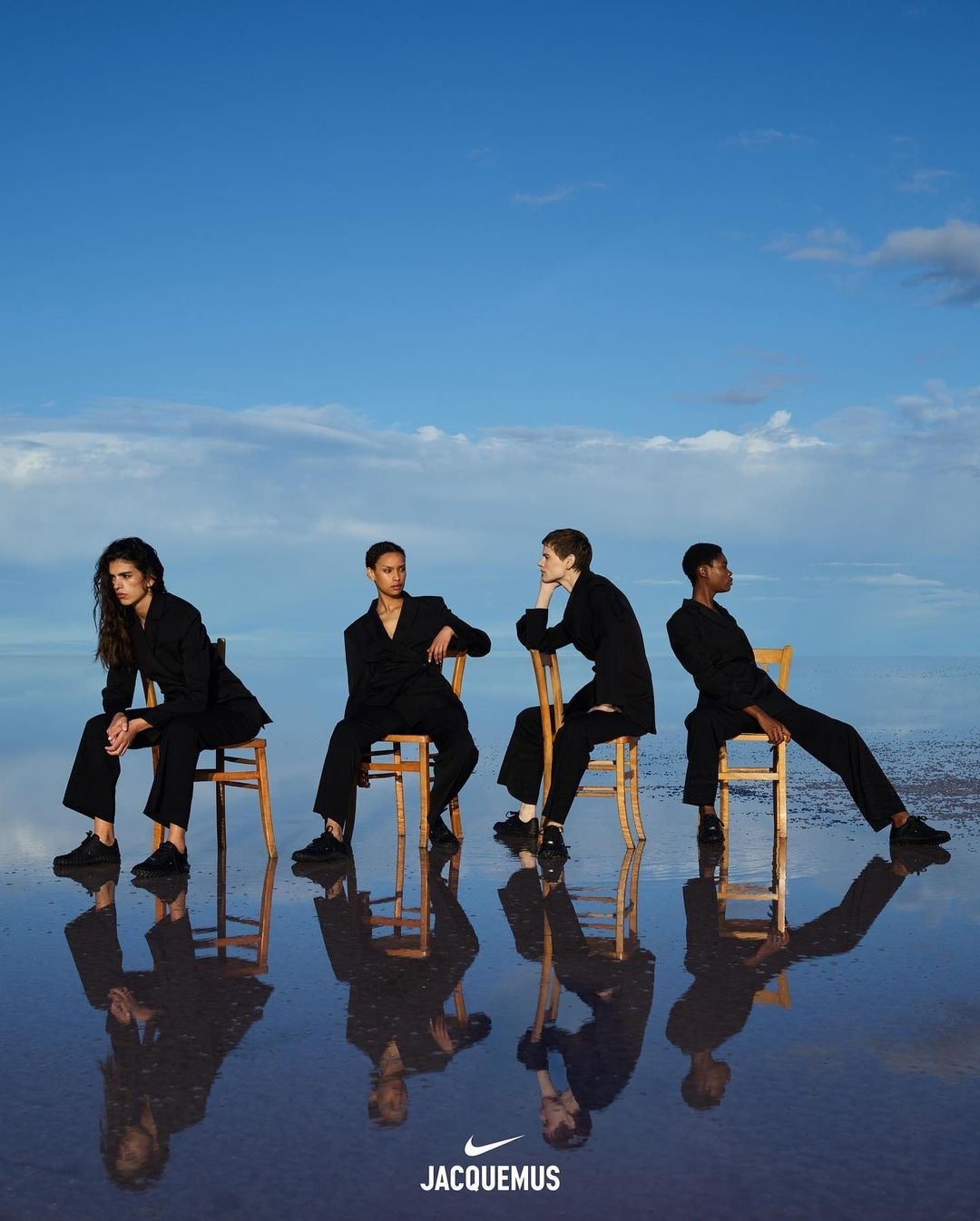 Four fashion models sit on chairs on a reflective water surface with a blue sky background, promoting Nike and Jacquemus.