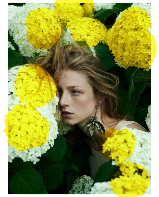 Blonde woman poses surrounded by yellow and white hydrangeas, wearing a leaf-shaped earring in a lush, verdant outdoor setting.