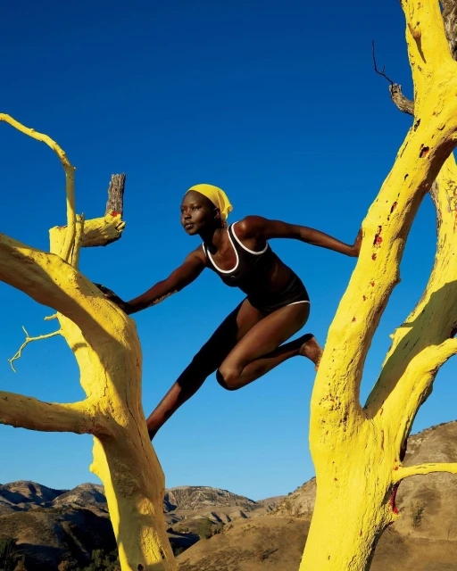 Woman in athletic swimsuit poses in a yellow painted tree against a blue sky and mountain landscape backdrop during daylight.