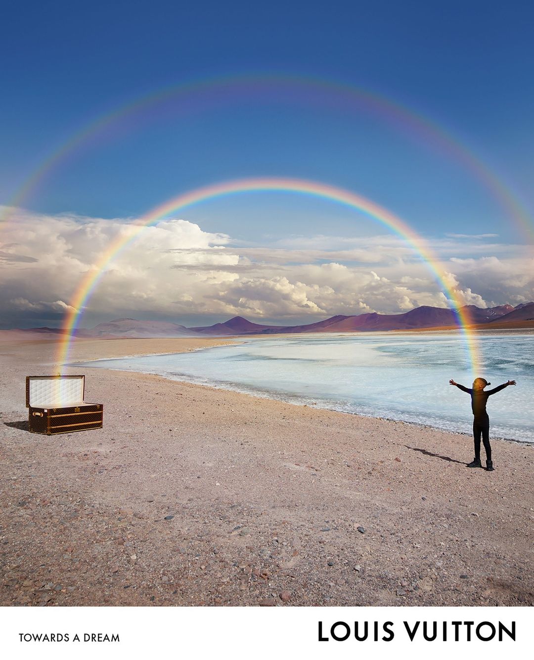 A person stands on a sandy beach by a calm body of water beneath a vibrant double rainbow, next to a vintage Louis Vuitton trunk.