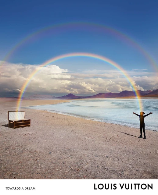 A person stands on a sandy beach by a calm body of water beneath a vibrant double rainbow, next to a vintage Louis Vuitton trunk.