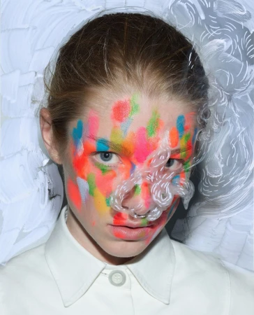 Close-up portrait of a model with vibrant multicolored paint on face and white collar shirt against a painted background