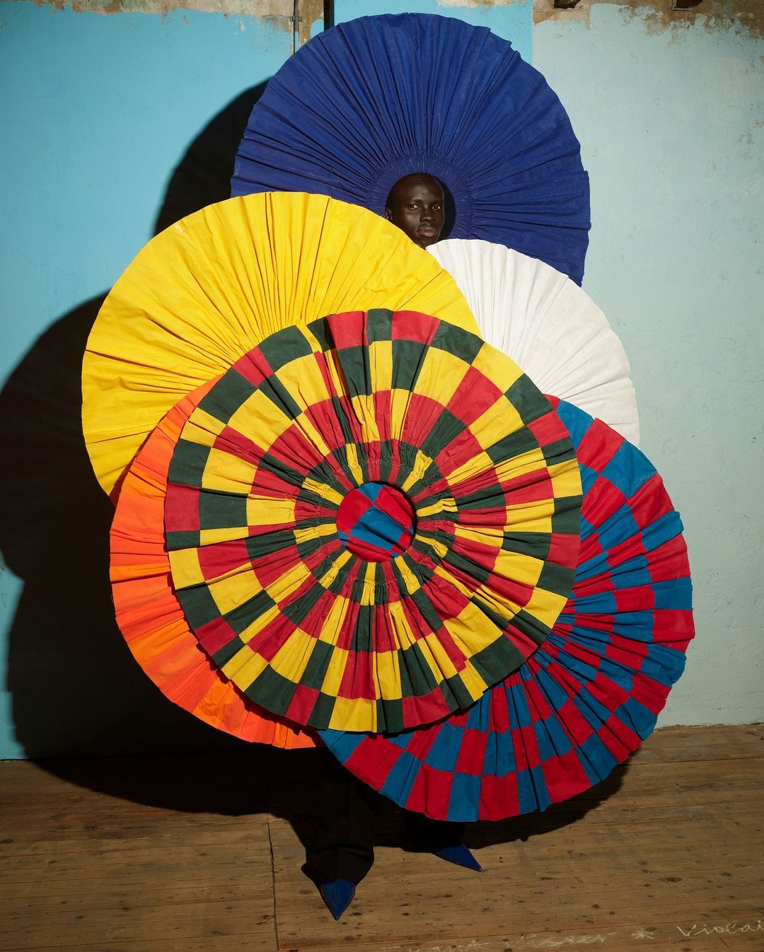 Person in surreal fashion costume of stacked, colorful, round textile forms against an old blue wall and aged wood flooring.