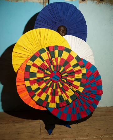 Person in surreal fashion costume of stacked, colorful, round textile forms against an old blue wall and aged wood flooring.