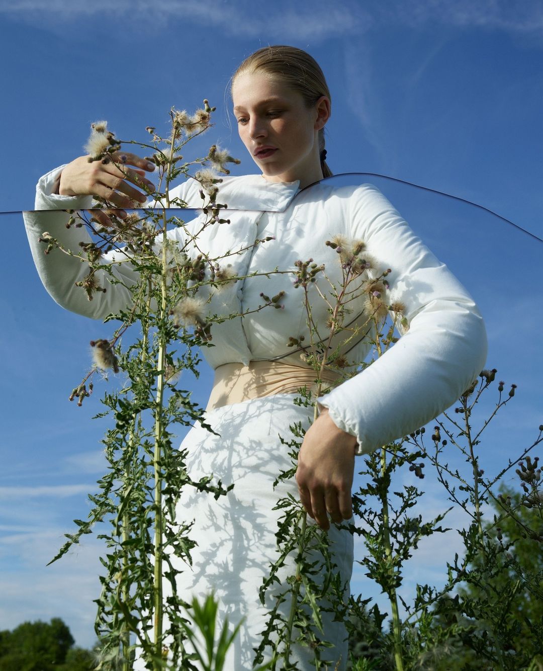 Blonde woman in white clothing poses among tall weeds under a blue sky in a fashion-style outdoor shot with creative glass filter effect.