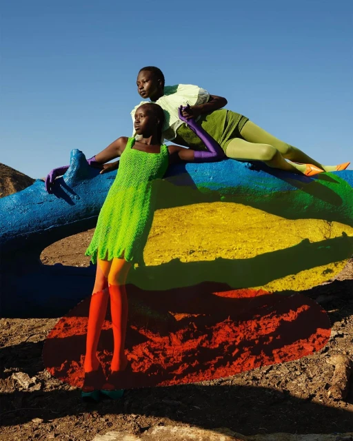 Two Black women in brightly colored outfits pose on rainbow painted surface in desert landscape with clear blue sky in editorial fashion image.