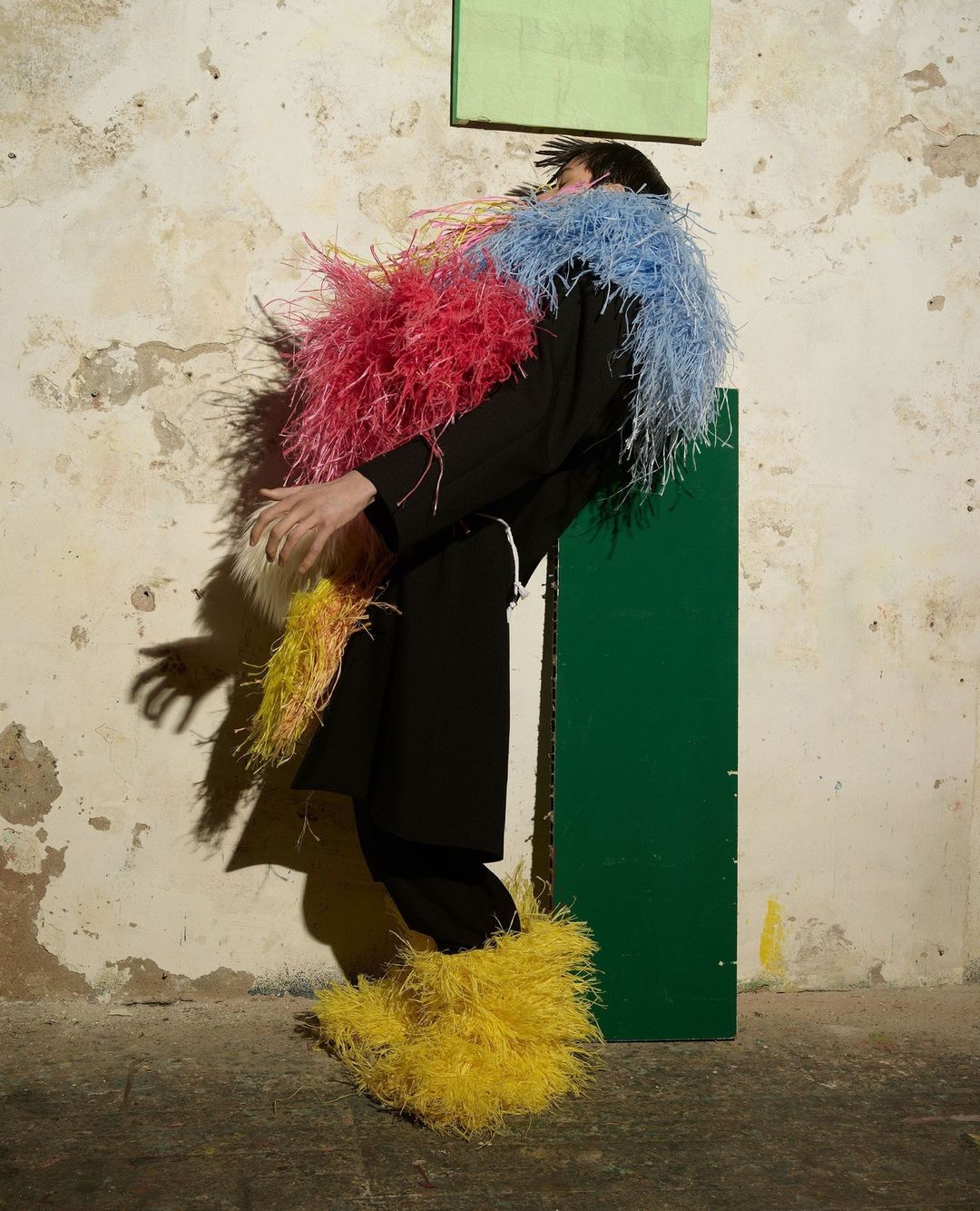 Person in black outfit with colorful feather boa and boot covers poses in front of a distressed wall and green/beige panels.