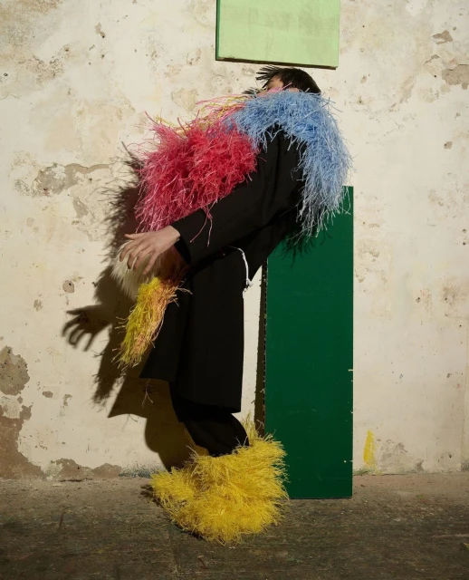 Person in black outfit with colorful feather boa and boot covers poses in front of a distressed wall and green/beige panels.