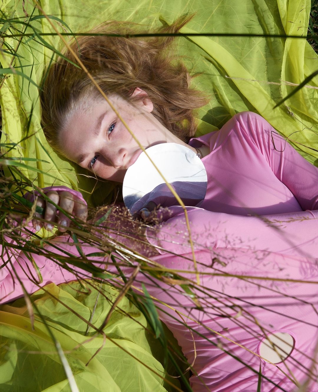 Young woman lies on green fabric in tall grass, holding a round mirror reflecting the sky. Fashion editorial, outdoor shot.