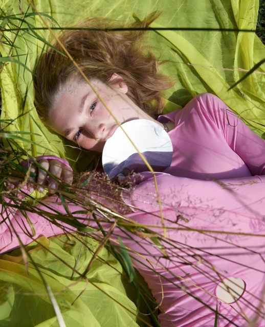 Young woman lies on green fabric in tall grass, holding a round mirror reflecting the sky. Fashion editorial, outdoor shot.