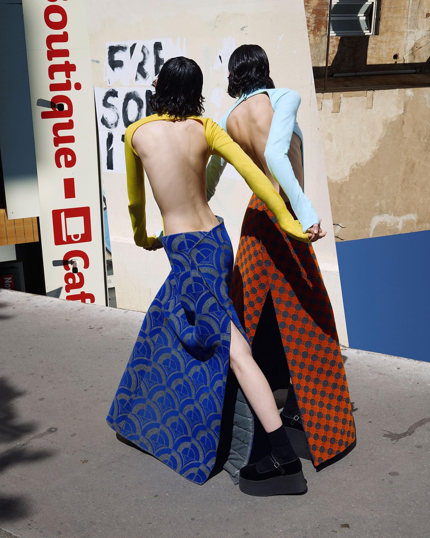 Two back-facing fashion models pose outdoors wearing long skirts with leg slits, long-sleeved tops, platform shoes, and dark bob haircuts.
