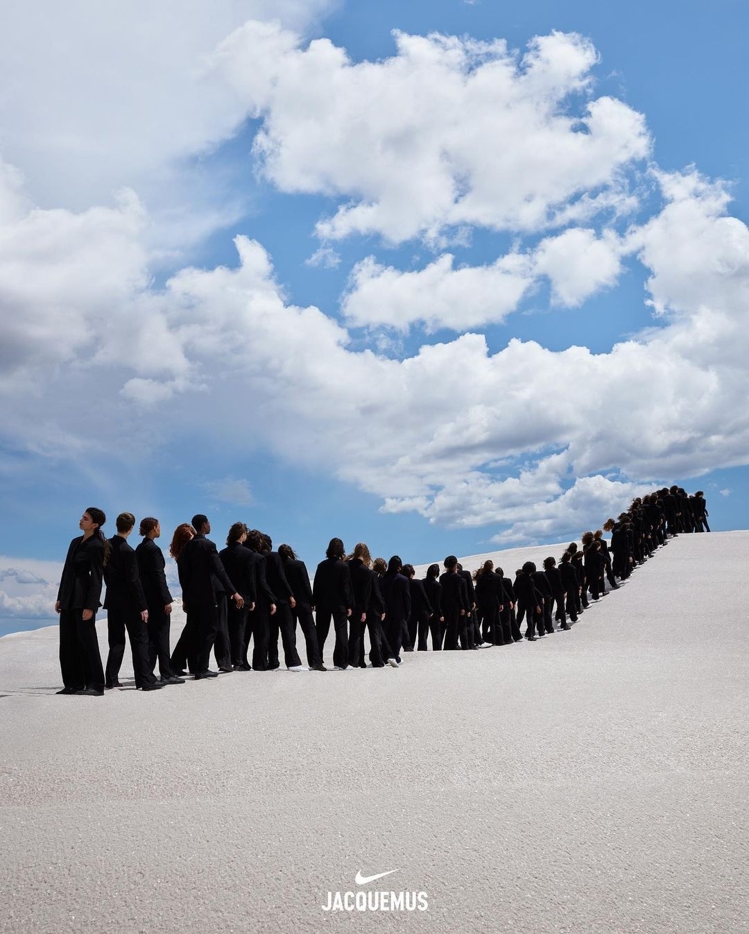 A large group of people in black clothing walking on sand under a blue, cloudy sky, in a fashion editorial shot with logo at the bottom.