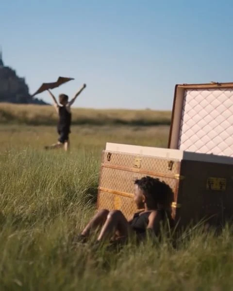 Woman lounges near a vintage trunk in a grassy field on a sunny day. In the background, a child runs with arms raised in a clear sky.