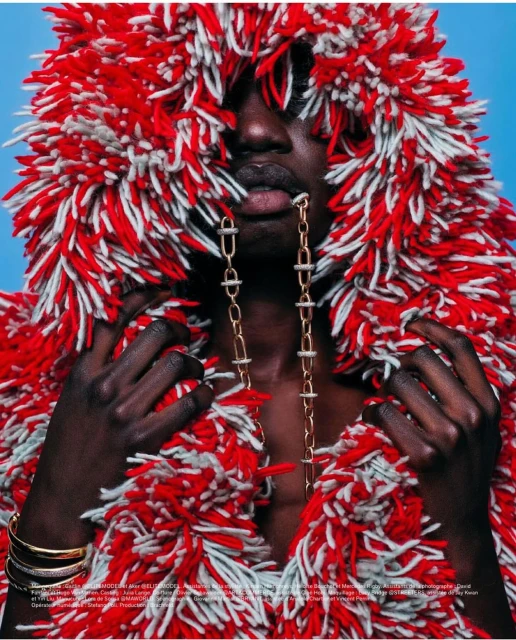 A striking portrait of a Black adult with a dramatic red and white textured hood, accessorized with a gold chain and bracelets, against a light blue backdrop.