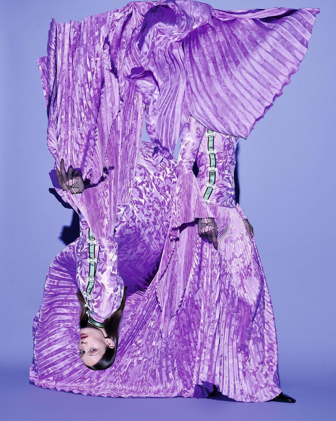 Fashion shot of a woman in a purple pleated gown with floral accents, upside down in front of a purple backdrop. The image is studio lit.