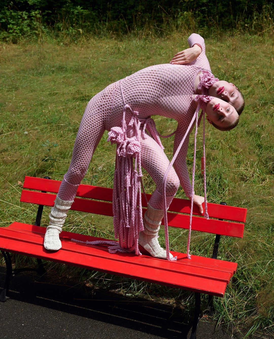 Two women in matching pink mesh outfits are bent over a red bench in a grassy park, with a sculptural crocheted accessory. 