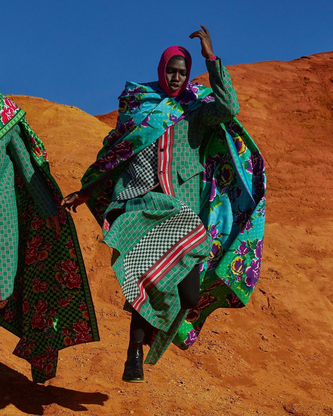 Fashion shot of a Black woman in a green floral cape and matching headscarf posing in a desert against a clear blue sky.