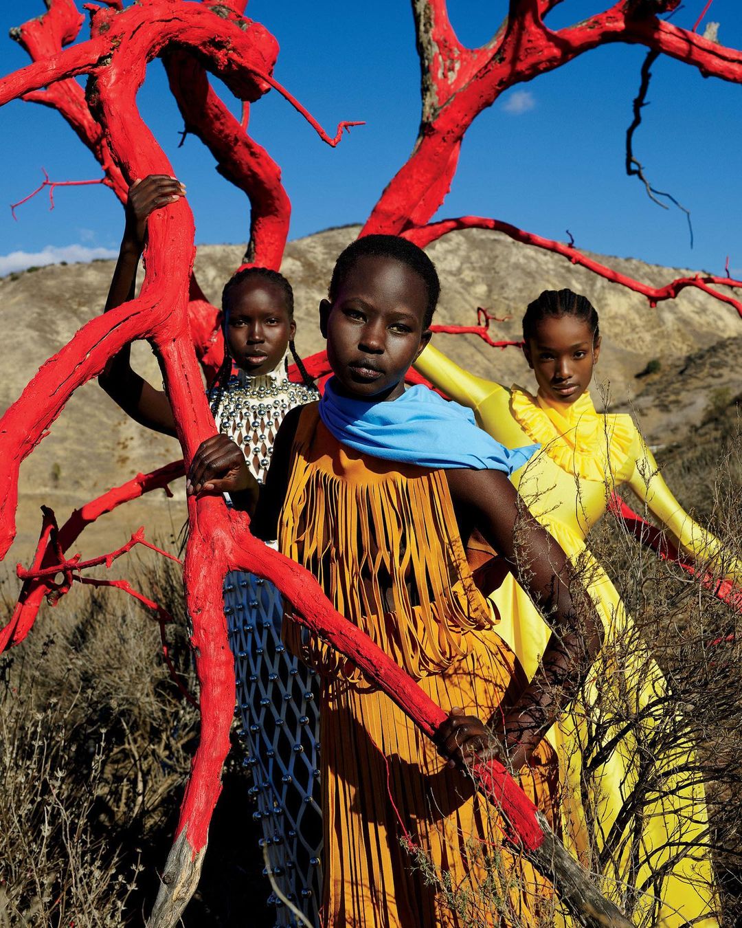 Three young women in colorful dresses pose amongst red-painted trees and arid hills under a bright blue sky. Fashion editorial photograph.