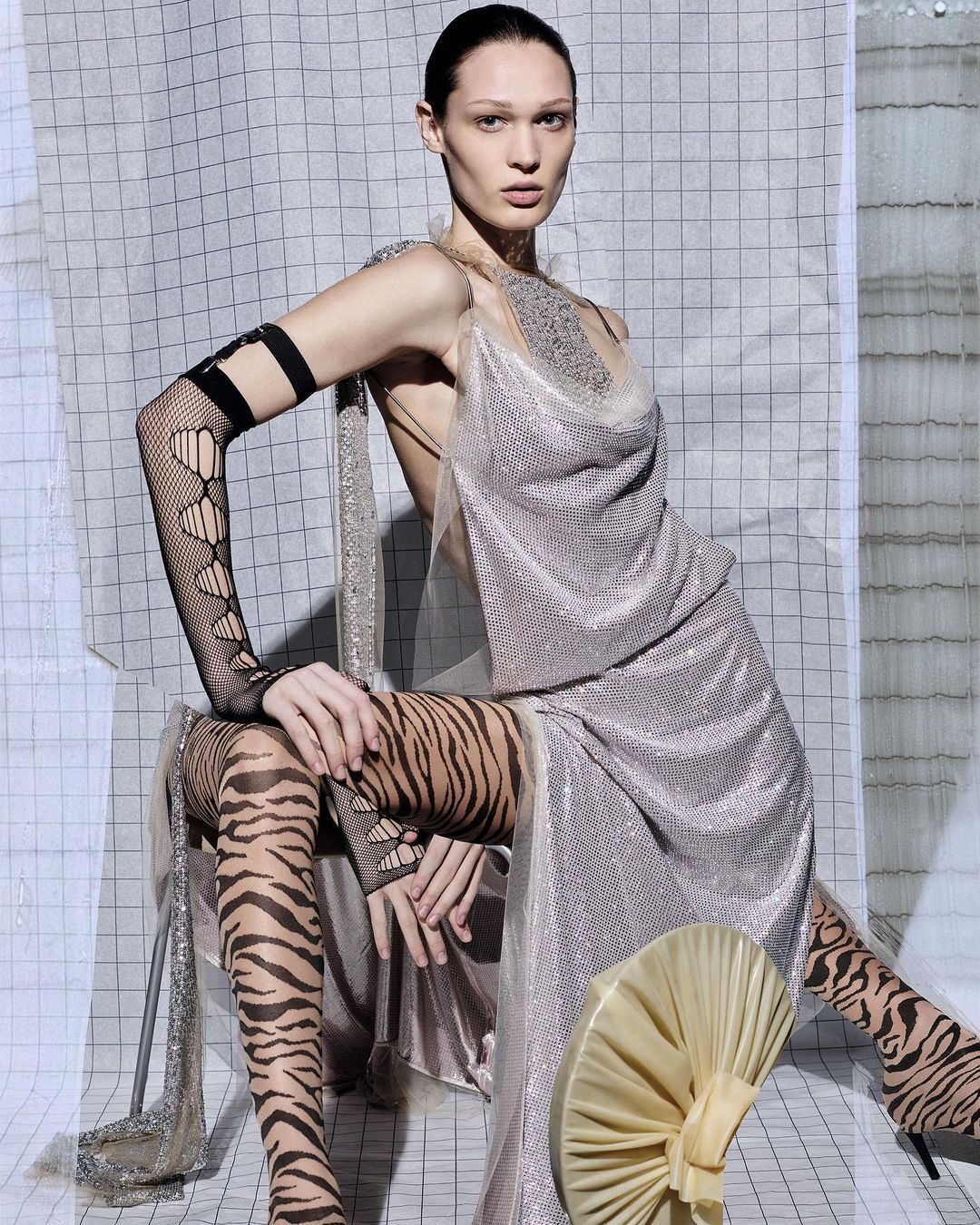 Adult female model in silver dress and tiger-print tights sits in front of a geometric patterned background in a fashion shot.