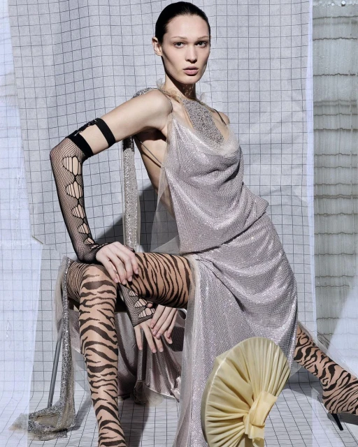 Adult female model in silver dress and tiger-print tights sits in front of a geometric patterned background in a fashion shot.