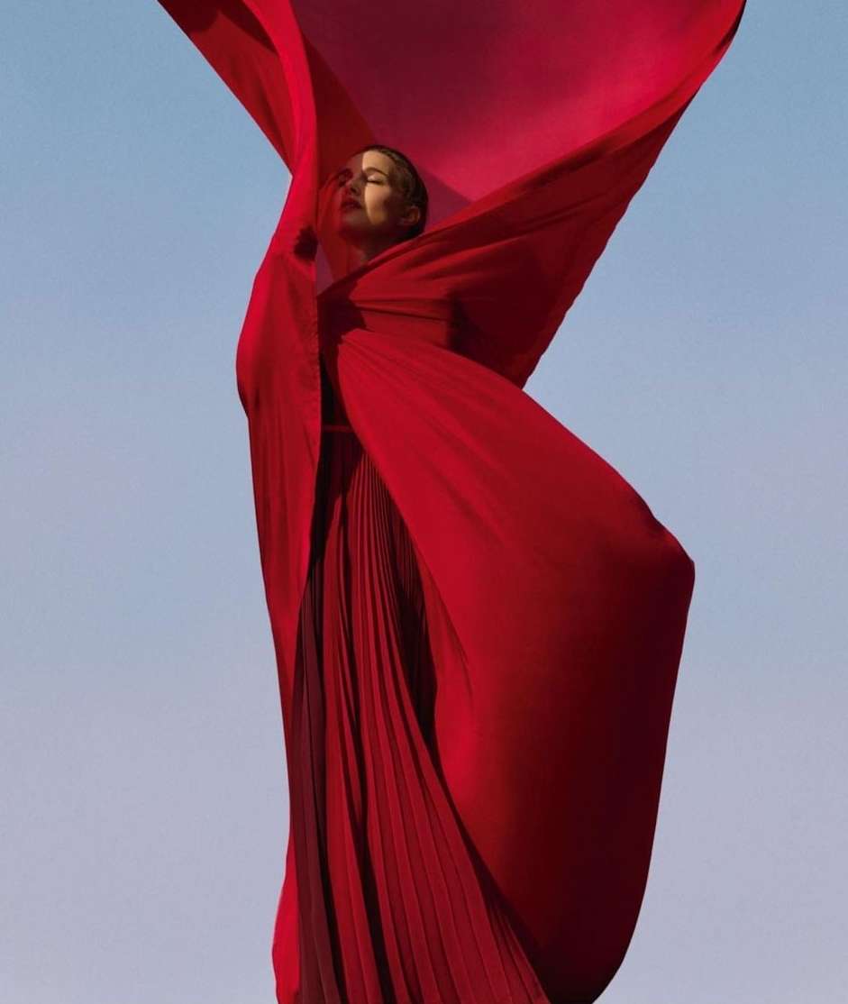 A woman in a long red dress stands against a pale blue sky, partially obscured by a flowing red fabric, in an eye-level shot.