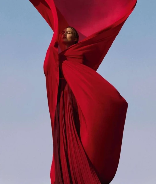 A woman in a long red dress stands against a pale blue sky, partially obscured by a flowing red fabric, in an eye-level shot.