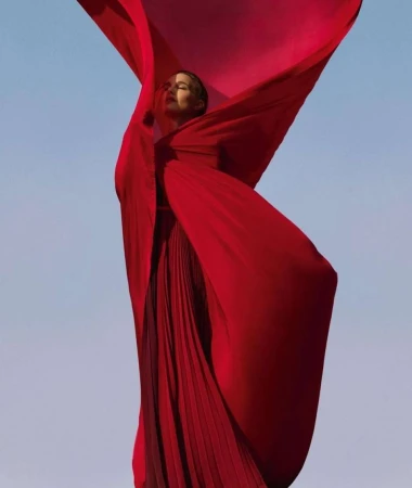 A woman in a long red dress stands against a pale blue sky, partially obscured by a flowing red fabric, in an eye-level shot.