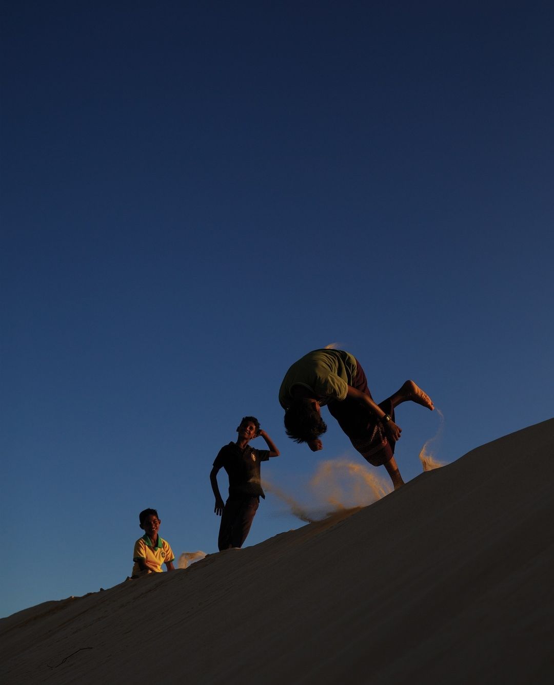 Three young people silhouetted against a blue sky on a sandy hill, with one doing a flip and kicking up sand.