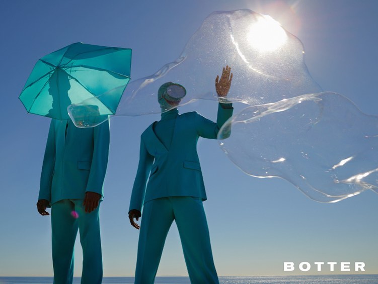 Two figures in teal suits, one with an umbrella for a head, stand on a beach under a bright sky, fashion-forward and surreal.