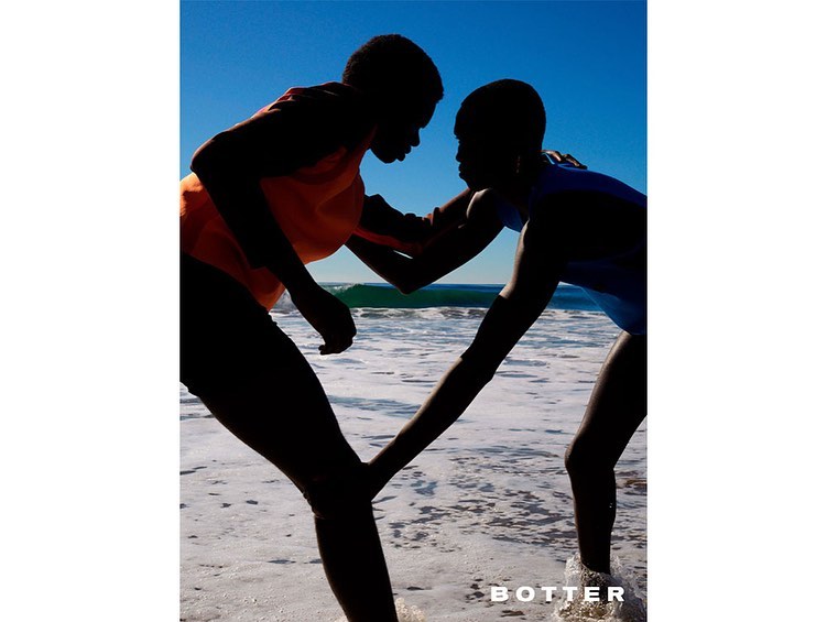 Two silhouetted figures in orange and blue swimwear face each other on a beach with a wave and clear sky, showcasing fashion brand BOTTER.
