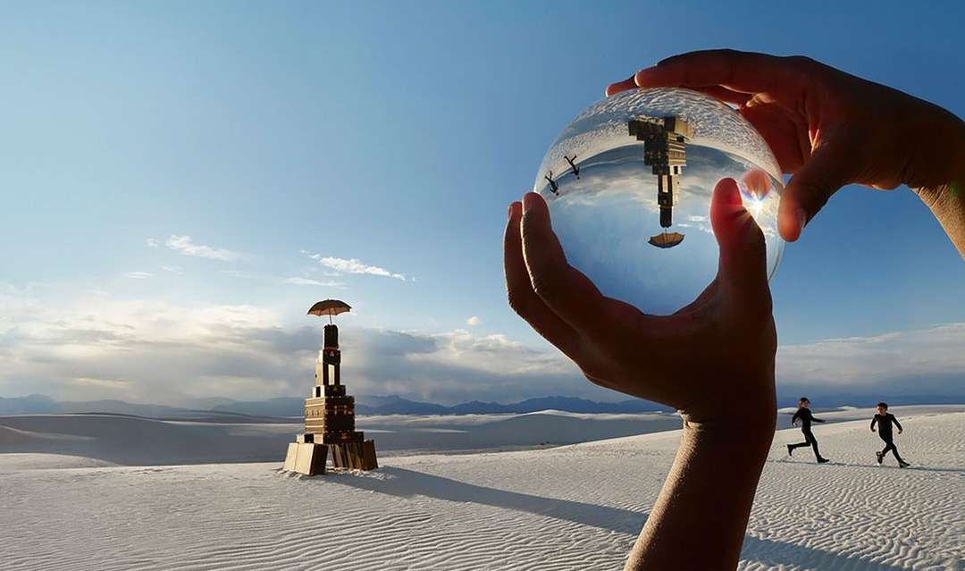 Artistic shot of sand dunes with a sculpture in a crystal ball, held by a person, under a blue sky with running people.