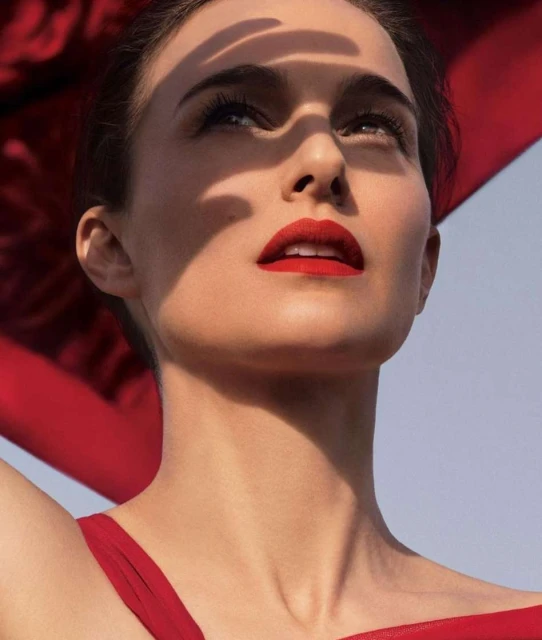 Portrait of a fair-skinned adult woman with red lipstick, looking up with a partly shadowed face, wearing a red top against a light blue sky.