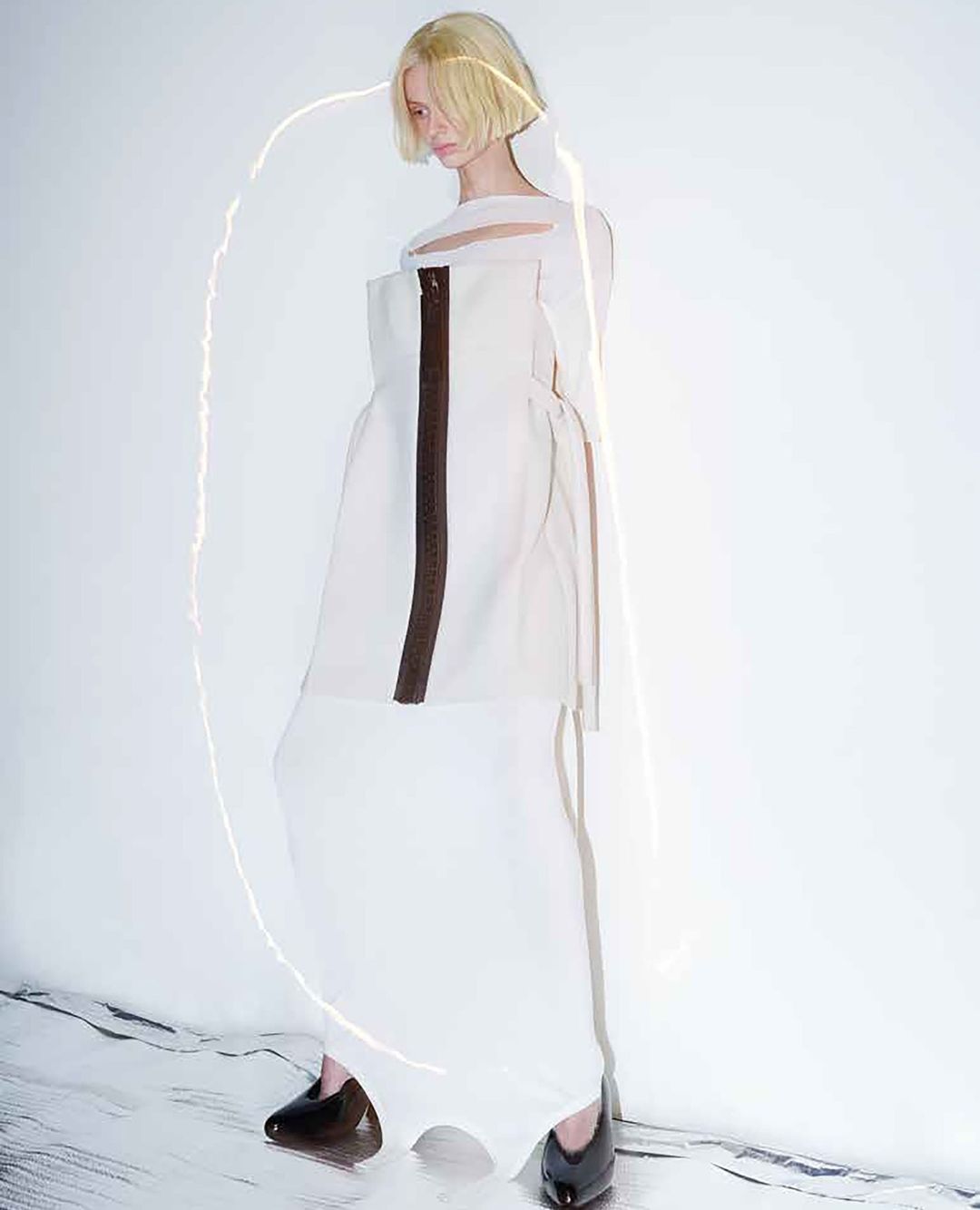 A solitary female fashion model in a sleeveless white dress poses against a bright, neutral background, illuminated by a curved light.