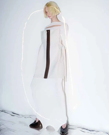 A solitary female fashion model in a sleeveless white dress poses against a bright, neutral background, illuminated by a curved light.