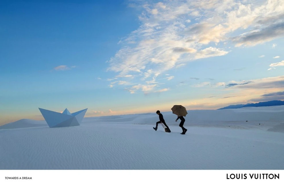 Paper origami boat on white sand dune with two silhouetted figures carrying a handbag and umbrella under a blue and cloudy sky.