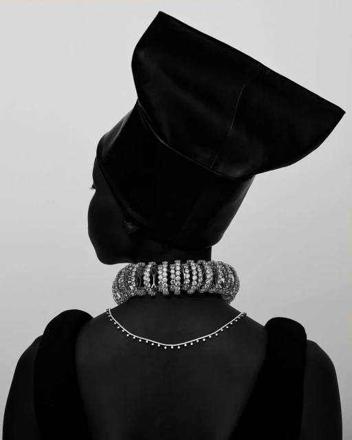 Back view of a woman with dark skin wearing a black headpiece and a jeweled choker necklace against a neutral background.
